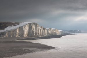 The snow-topped white chalk cliffs of Seven Sisters with a shingle beach in the bottom left, a grey sky behind and a calm sea. 