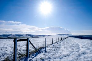 A long fence with a diminishing perspective sitting on the top of white snow covered mount Caburn, Lewes, East Sussex, United Kingdom, snow covered rolling hills are behind, a bright shining sun sits in the middle of a blue sky on a cold, clear winters day.