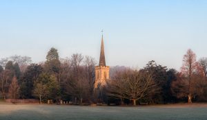 An old church with a tall spire in the middle of a frosted field with a line of trees behind and a bright blue sky 