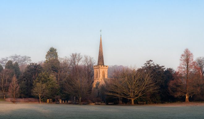 An old church with a tall spire in the middle of a frosted field with a line of trees behind and a bright blue sky