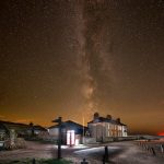 The milky way rises over a cottage by a cliff