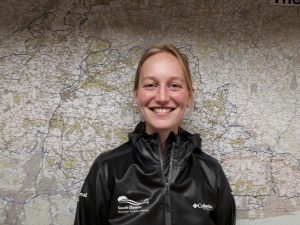 A blonde haired woman in a black coat which says 'South Downs National Park Authority' smiles at the camera in front of a map of the South Downs.